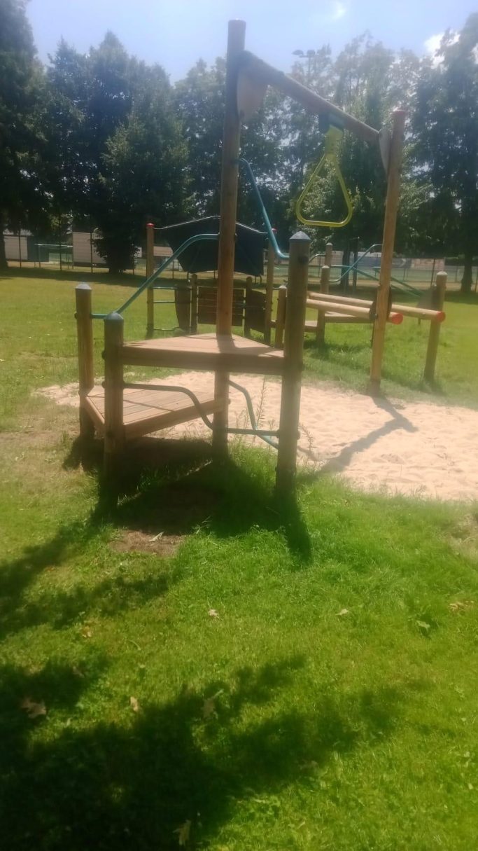 Holzspielplatz mit Klettergerüst und Rasenfläche im Hintergrund.